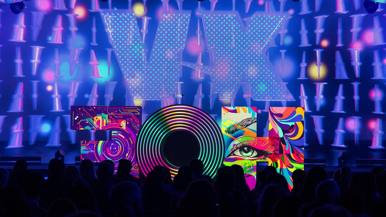 A large sculpture of the letters “HOF” sits atop the 满帆 Live Venue stage with different multicolored images projected onto each letter. The letters are in front of a large purple screen featuring the Roman numeral “XV” in large font. In front of the stage is a black silhouette of the crowd.